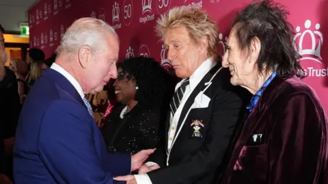 PA Media The King greets Sir Rod, who is dressed in a black blazer, white shirt and black and white striped tie. To the left of the image is Ronnie Wood, who is in a purple velvet jacket with a light blue shirt underneath which features a butterfly design on it
