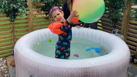 A boy in a round pool of water wearing a swimming outfit covered in dinosaurs, goggles and red water wings holding a large ball