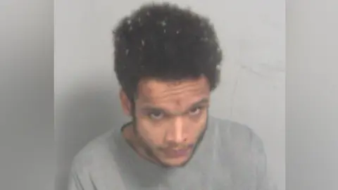 Essex Police A man with a short beard and moustache and tall brown hair sits in a grey long-sleeve jumper looking at the camera 