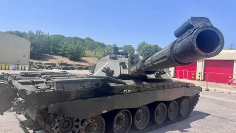 A Challenger 2 is parked before red garages under a bright blue sky. It's gun extends towards the right of camera. Behind the tank are service vehicles and trees dot the skyline. 