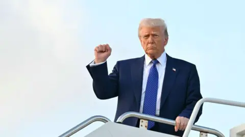 Getty Images Donald Trump raises his fist in the air while at the top of some stairs leading from a plane, and he holds the railing