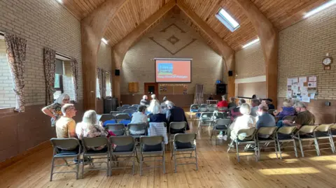 A number of people are seated in a hall and are looking at an overhead screen which says "Keep Wind Off Woldgate". 