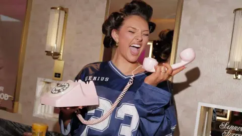 Tatty Lomas A women wearing a blue football jersey, with her hair in rollers, holds a vintage-style pink phone.