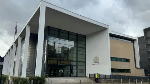 George King/BBC The front of Ipswich Crown Court, a modern building with a huge multi-storey glass facade. A person in a high-visibility jacket is standing outside with their back turned.