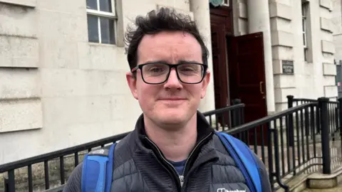 Dr Pádraig Ó Tiarnaigh looking at the camera. Pádraig has dark brown hair and is wearing a black jacket and glasses.