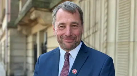 LCC An image of Councillor Stephen Atkinson standing in front of County Hall. He is wearing a navy suit and white shirt with a red tie and a lapel badge of the Lancashire red rose.