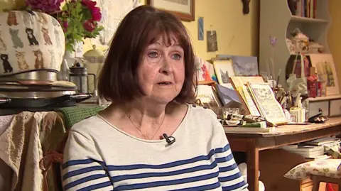 BBC Carol Smithers with red, short hair, wearing a white and blue striped long-sleeved top, sits in a chair in a house.