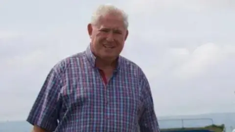 Andrew Hodson has short grey hair and is wearing a purple and blue checked shirt. He is stood on the coast in Wirral with the sea in the background.