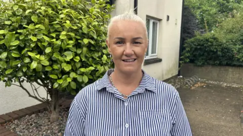 A woman with white hair smiles at the camera. She is wearing a navy and white stripy shirt. In the background you can see the side of a white house with a green tree the right of the photo