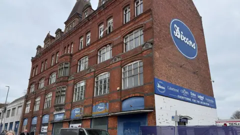 A five-storey building photographed at an angle. The Dixons logo, white words on a blue background, is in various places, with the largest version on the side of the building.