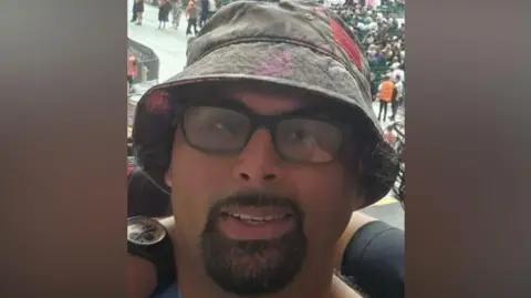 Police handout Lakhbir is wearing a grey bucket hat and glasses and smiling at the camera. He appears to be at a busy outdoor event as people can be seen in the distance behind him.
