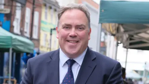 Jason Perry, wearing a navy suit and a spotty tie, smiling at an outdoor market