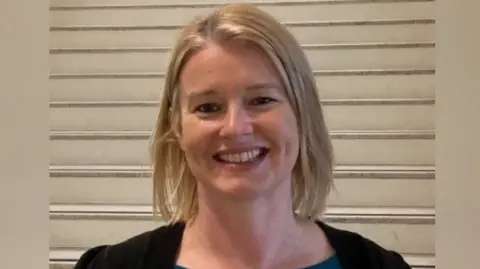 Anne Madden Anne Madden - a woman with shoulder-length blonde hair smiles at the camera. She is wearing a black cardigan and is standing in front of white stairs.