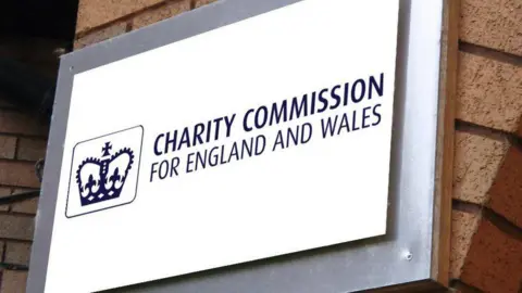 Blue and white sign showing charity commission building. The sign has a crown and is which hung on a brick pillar.