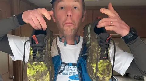 Max Howard The picture shows a man holding up a pair of very muddy running shoes. The shoes are covered in thick dirt, especially around the toes and sides, suggesting they have been heavily used on wet or rough terrain.