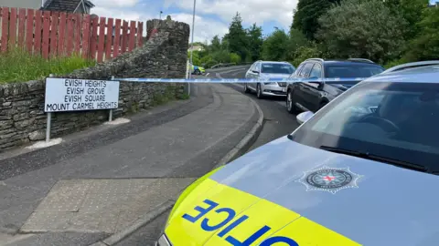 a police car is parked in front of a police cordon. a number of homes can be seen in the background, as can a number of parked cars
