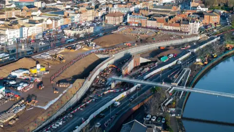 Stockton-on-Tees Borough Council An aerial view of a narrow, modern white footbridge over the River Tees. The bend of the river can be seen to the right with the bridge disappearing out of shot. On the left, the river gives way to yellow building machinery on the bank. In the foreground is a building with a tiled roof behind which a line of cars are parked. A road along the bank has tall silver fencing on one side and short red and white barriers on the other. There is a large expanse of flattened earth between the road and the buildings of Stockton.