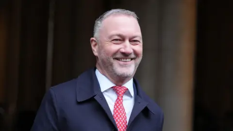 PA Media A man in his fifties smiling with short white hair and a trimmed white beard. He is walking and wearing a white shirt, red tie and dark blue mackintosh coat.