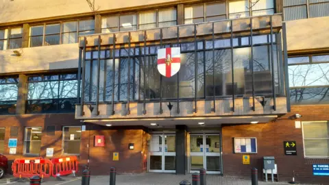 Stock photo of City of Lincoln Council building on Orchard Street. The building has large windows and a mix of brick and concrete. Above the entrance is a shield with a red cross and an emblem, surrounded by a black metal frame. The entrance has glass double doors and is surrounded by paved ground with black bollards in front. To the left are red maintenance barriers.
