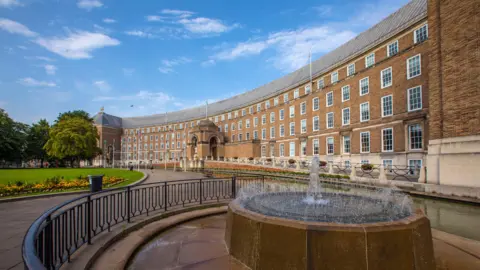 Getty Images Bristol City Council Town Hall. It is a wide, brown brick building with lots of windows. There are flagpoles at the front of the building and you can also see a fountain. 