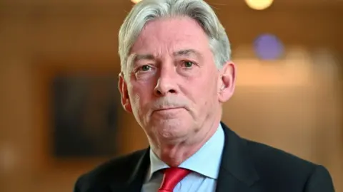 Getty Images A man with grey hair, swept back over his head, looks towards the camera in the Scottish Parliament. He is wearing a black suit and red tie with a light blue shirt. 