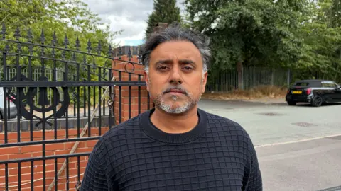 Manraj Sanghera standing outside Park Street South in Blakenhall. He is in front of a red brick wall and gate. He has short black hair with hints of grey and a white and grey beard. He wears a black jumper.