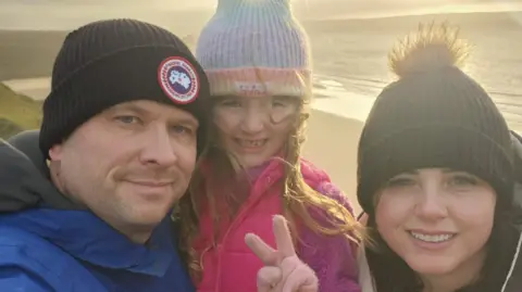 Angharad Dennis Angharad Dennis smiles as she takes a selfie with her husband and daughter on a beach. They wear hats and coats with the sun behind them.