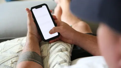 A man with a tattoo on his arm looks at a white mobile phone screen