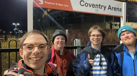 Four young men at Coventry Arena Railway Station. Stood in front of the station sign on the platform is a man with yellow and purple glasses, a man with red coat, glasses and black hat, a man wearing glasses with a blue coat with blue and white striped top, and a man with a blue jacket on, ginger beard and turquoise beanie hat 