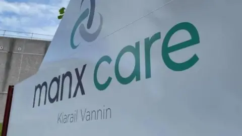 A close-up of a white sign that reads Manx Care in grey and green lettering, with the Manx translation underneath.
