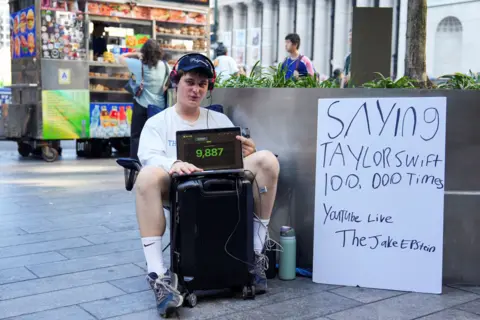 Reuters A young man sits on the street outside the venue holding a laptop with the number '9,887' on screen, and a placard next to him reading 'saying Taylor Swift 100,000 times, YouTube live'.