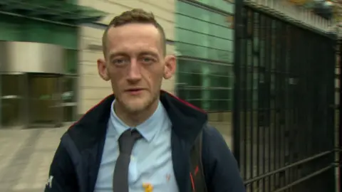 Craig Rowland has short fair hair and has stubble on his face.
He is wearing a blue shirt an grey tie and is seen outside Laganside Court in Belfast.