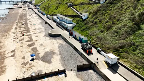Serco/NNDC Ariel view of Cromer beach with the pier in the distance. The grass covered dunes end at a promenade lined with beach huts. A refuse lorry and staff wearing high-vis jackets are on the promenade. People are on the beach and in the sea. 