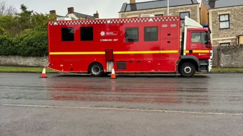 A red fire engine in front of some houses on a road. There are two traffic cones towards the back of the fire engine.