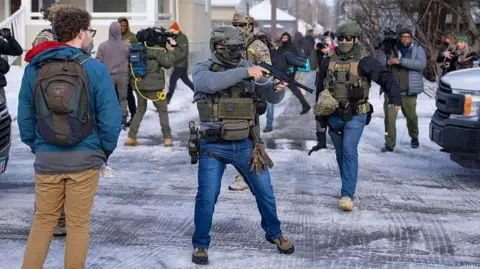 An officer with US Immigration and Customs Enforcement or ICE (right) and another federal officer holding a crowd control device (centre) stand at a Minneapolis intersection where protesters had gathered after the death of Renee Nicole Good. Bystanders and journalists also stand in the intersection.