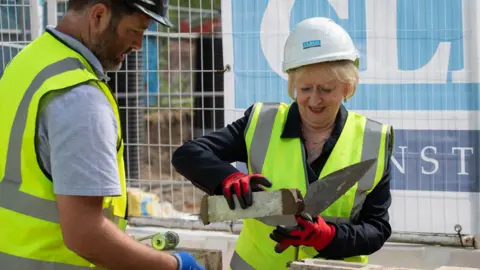 A woman with short blonde hair lays a brick wearing a high-vis yellow vest, hard hat and gloves. A man, also insafety gear, assists her. 