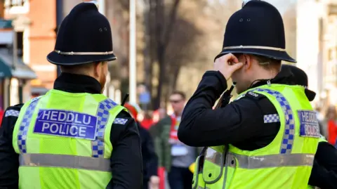 Two police officers wearing yellow high visibility jackets with "Heddlu" and "Police" written on the back.