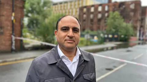 Yunus Mulla/BBC Abdullah Alumair, who has receding black hair and wears a black leather jacket over a white shirt, is pictured with the charred remains of the Hotspur Press building in the background