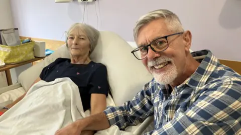 A woman with short grey hair and a blue top lies in a hospital bed next to a man, with a blue and white shirt and wearing glasses, sits in a chair next to her.