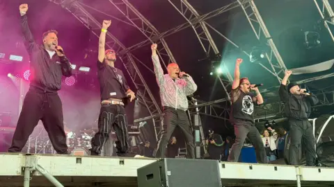 Boy band Five, standing in a line on stage - all singing with their right arms in a fist in the air