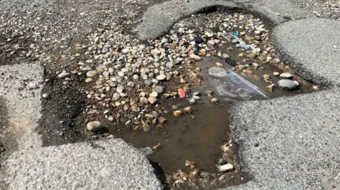 A pothole in a road.  Pebbles and water along with a plastic bag fill the hole in the middle of some tarmac.