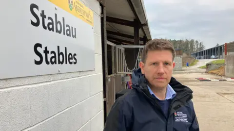 A photo of lead researcher Dr Sebsatian McBride. There is a sign that reads stables to the left of him in English and Welsh. He is wearing a blue jacket and is looking at the camera.