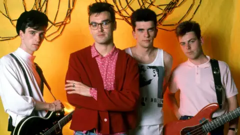 Getty Images The Smiths' guitarist Johnny Marr, in a white shirt, singer Morrissey, in glasses and a pink and white spotted shirt and a red cardigan, drummer Mike Joyce, in a white vest, and bassist Andy Rourke, in a pink polo shirt pose for a portrait before their first show in Detroit during the 1985 Meat Is Murder Tour 