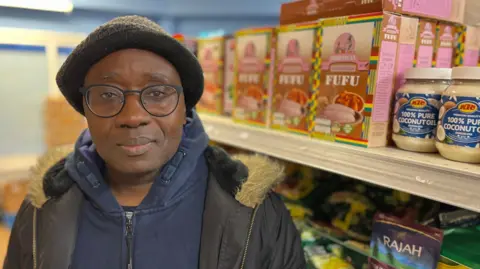 Olusegun Adedoyin standing in front of a rack of jars and packets in his store Olly Faith on Saint Luke's Terrace. He's wearing a hat and glasses and has on a black puffer coat over a dark blue fleece.