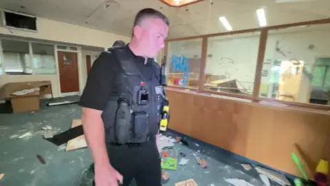 A police officer walking through a building that appears top have been vandalised