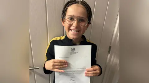 A boy with braided hair and large glasses smiles as he holds up a letter from Downing Street. 