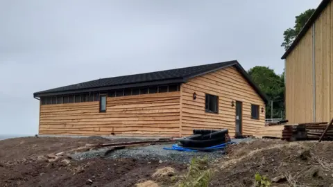 A wooden lodge with a pitched roof is built on land with bits of pipe dumped next to the structure. 