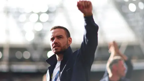 Getty Images Liam Payne holding his fist in the air standing in a football stadium. He is wearing a blue jacket, a white t shirt and a lanyard sits around his neck.