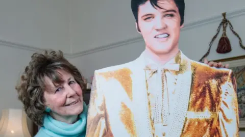 Anne Murphy Anne Murphey - a woman with dark hair and a turquoise jumper, holding cardboard cut out of Elvis Presley.