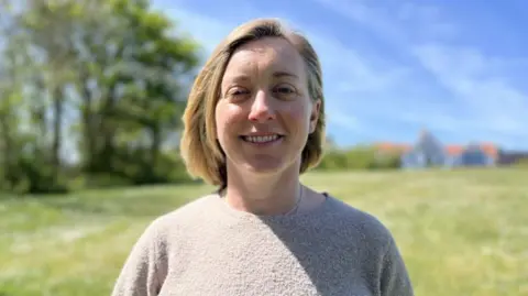 Charlie Cox is smiling at the camera as she stands in a sunny field. She has shoulder-length blonde hair and blue eyes., She is wearing a grey sweater.